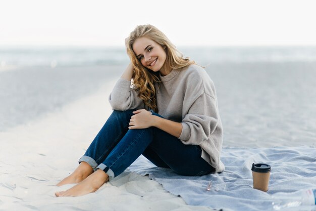 Encantadora mujer caucásica escalofriante en la playa de otoño. Increíble modelo femenino rizado disfrutando de un café en la arena.