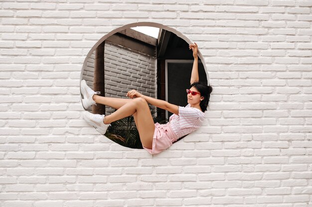 Encantadora mujer caucásica sentada en la pared de ladrillos. Tiro al aire libre de mujer bronceada alegre posando sobre fondo urbano.