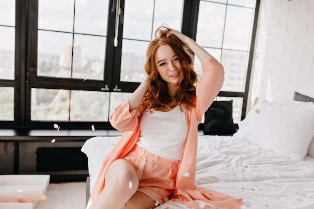 Encantadora mujer caucásica tocando su cabello ondulado mientras posa en la mañana. Sonriente niña complacida en ropa de dormir disfrutando de fin de semana.