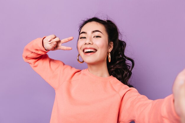 Encantadora mujer coreana posando con el signo de la paz. Riendo a joven asiática tomando selfie sobre fondo púrpura.