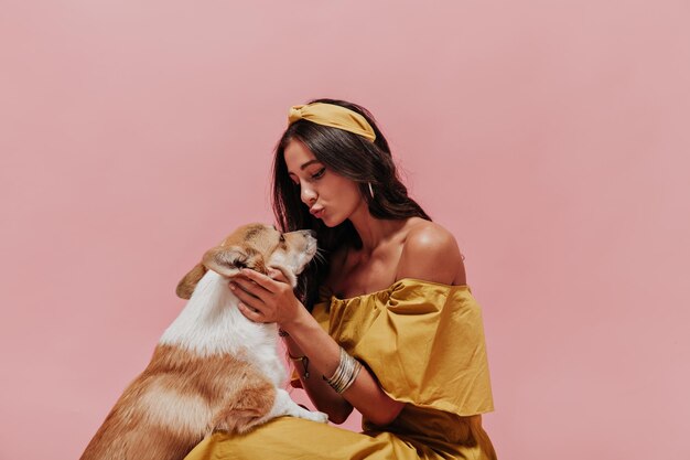 Encantadora mujer delgada con estilo con el pelo largo y oscuro en bralettes drendy y traje amarillo jugando con un pequeño corgi sobre fondo rosa