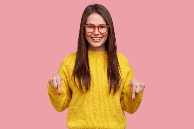 Encantadora mujer despreocupada con expresión positiva, apunta hacia abajo con ambos dedos índices, vestida con ropa informal, tiene una amplia sonrisa interesada