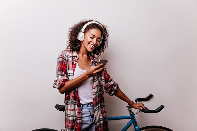 Encantadora mujer elegante en auriculares leyendo mensajes telefónicos. Chica africana bien vestida con bicicleta con smartphone.