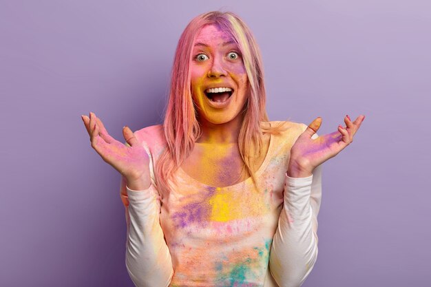 Encantadora mujer emocionada se ríe alegremente, levanta la mano, tiene el rostro multicolor cubierto con pintura en polvo durante el festival de Holi, celebra la fiesta india y la llegada de la primavera, modelos sobre una pared púrpura