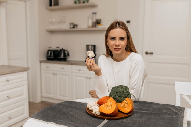 Encantadora mujer encantadora está posando en la cámara mientras está sentado en la cocina con una mesa de verduras