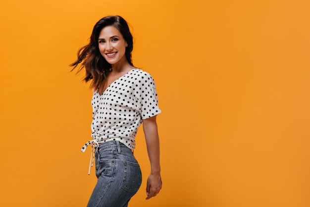 Encantadora mujer se enciende sobre fondo naranja. Linda chica de buen humor con pelo corto en camisa de lunares y jeans azul claro está sonriendo.