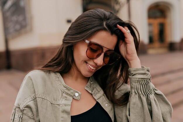 Encantadora mujer europea tímida con cabello oscuro con gafas de sol con chaqueta caminando por la ciudad en buen día soleado