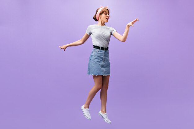 Encantadora mujer en falda de mezclilla salta sobre fondo morado. Chica guapa de moda en diadema rosa y camisa gris posando.