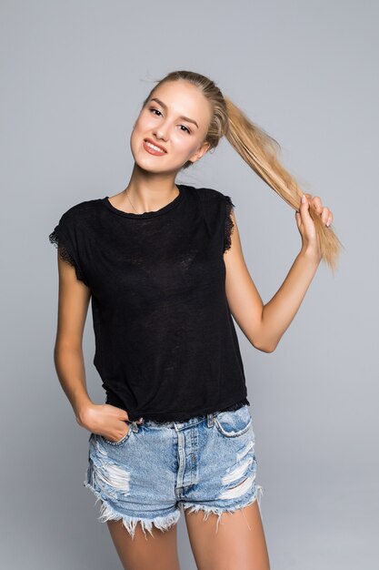 Encantadora mujer feliz encantadora viste camiseta blanca y pantalones cortos de mezclilla sonriendo a la cámara y tocando su cabello sobre fondo gris aislado