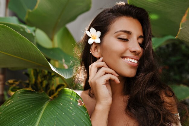 Encantadora mujer con flor blanca en cabello oscuro sonríe dulcemente con los ojos cerrados entre árboles tropicales con hojas grandes