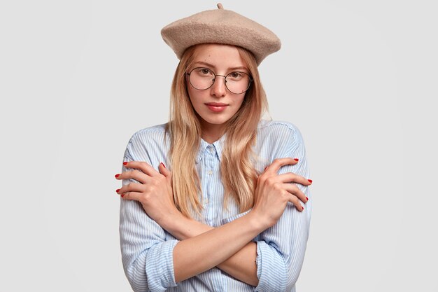 Encantadora mujer francesa con elegante boina y camisa, mantiene las manos cruzadas, ha reservado la expresión facial, aislada sobre una pared blanca. Hermosa mujer tímida en gafas se encuentra en el interior. Belleza y juventud