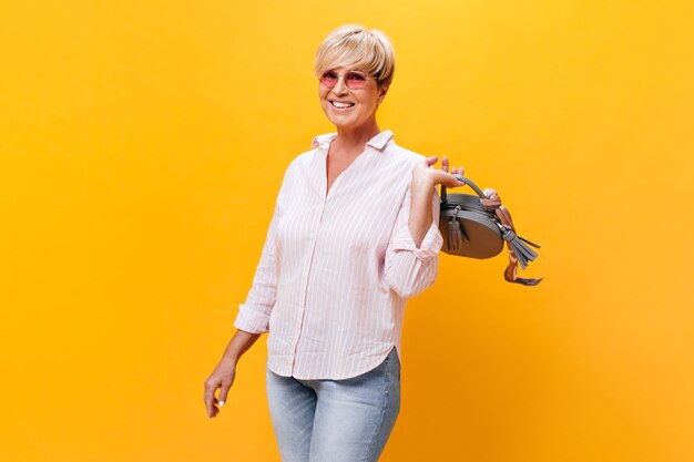 Encantadora mujer con gafas de sol y camisa rosa posando con bolso sobre fondo naranja