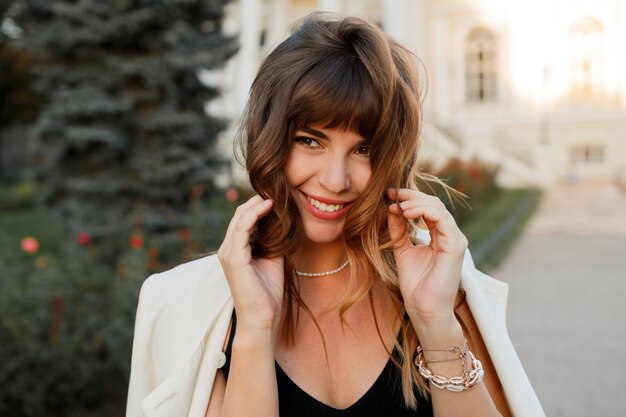 Encantadora mujer hermosa con peinado ondulado sonriendo, coqueteando, estado de ánimo romántico. Vistiendo chaqueta blanca. Al aire libre. Moda de otoño.