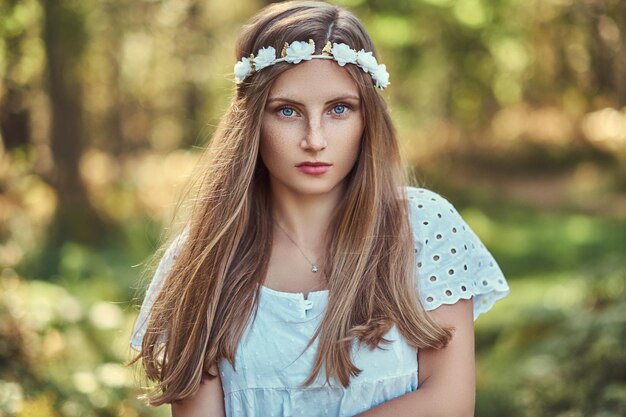 Una encantadora mujer hermosa con un vestido blanco y una corona blanca en la cabeza posando en un bosque verde de otoño.