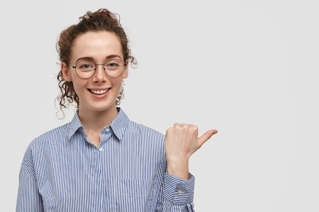 Foto gratuita encantadora mujer joven atractiva pecosa con expresión alegre, tiene pecas en la piel, vestida con elegante camisa, apunta a un lado con el pulgar