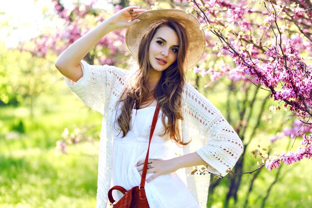 Encantadora mujer joven y bonita con pelo largo con sombrero de verano, vestido de luz blanca caminando en el soleado jardín sobre fondo floreciente de sakura. Relajación, sonreír a la cámara, ropa ligera, sensible, alegría