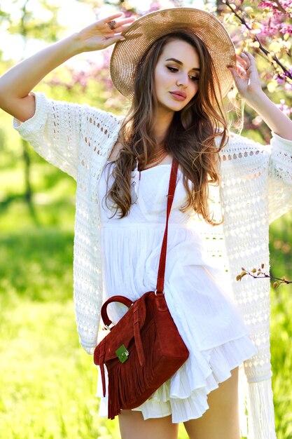 Encantadora mujer joven y bonita con pelo largo con sombrero de verano, vestido de luz blanca caminando en el soleado jardín sobre fondo floreciente de sakura. Relajación, sonreír a la cámara, ropa ligera, sensible, alegría