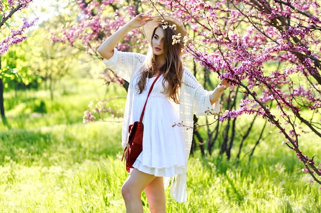 Encantadora mujer joven y bonita con pelo largo con sombrero de verano, vestido de luz blanca caminando en el soleado jardín sobre fondo floreciente de sakura. Relajación, sonreír a la cámara, ropa ligera, sensible, alegría