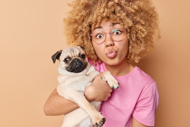 Encantadora mujer joven de cabello rizado mantiene los labios plegados abraza al perro pug tiene un nuevo miembro de la familia que usa anteojos transparentes y camiseta aislada sobre fondo beige Personas y animales domésticos