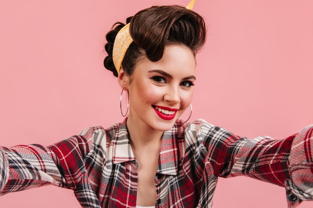 Foto gratuita encantadora mujer joven en camisa a cuadros sonriendo sobre fondo rosa. chica pinup emocional tomando selfie.