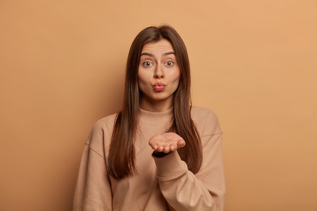 Encantadora mujer joven y cariñosa tiene una piel sana, mantiene los labios doblados, sopla un beso al aire, te envía mwah, tiene la palma de la mano extendida, cabello largo y oscuro