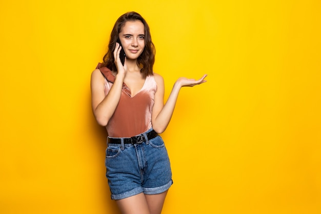 Encantadora mujer joven contenido sorprendido hablando por teléfono celular sobre pared amarilla