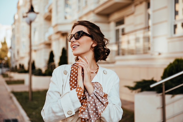 Encantadora mujer joven en elegantes gafas de sol disfrutando de un cálido día de otoño. Tiro al aire libre de chica rizada alegre en chaqueta blanca.