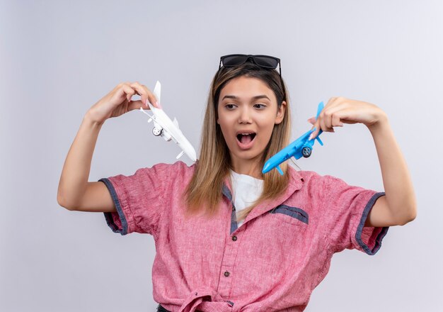 Una encantadora mujer joven emocionada con camisa roja con gafas de sol mirando sorprendentemente aviones de juguete blancos y azules mientras los volaba