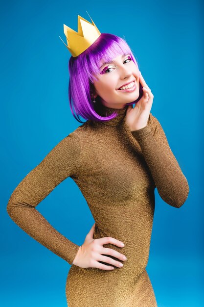 Encantadora mujer joven con estilo en vestido de lujo dorado sonriendo. Corta el cabello morado, corona, sonrisa amistosa, expresando positividad, felicidad, celebrando el carnaval, fiesta.