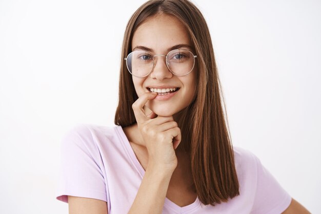 Encantadora mujer joven femenina coqueta y segura de gafas sosteniendo el dedo en el labio inferior y sonriendo con deseo