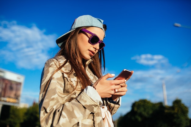 Encantadora mujer joven en gafas de sol usando un teléfono móvil, navegar por los sitios web