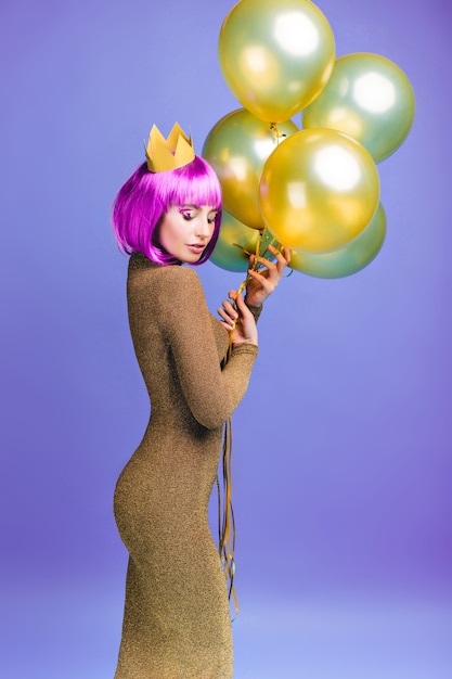 Encantadora mujer joven hermosa en atractivo vestido de moda con globos dorados voladores. Corte de pelo rosa púrpura, corona, emociones alegres, ojos cerrados, celebración.