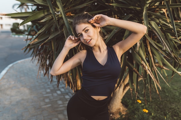 Encantadora mujer joven increíble en ropa deportiva sonriendo en árbol tropical. Estirar las manos, figura sexy, modelo deportivo, humor alegre, verdaderas emociones positivas