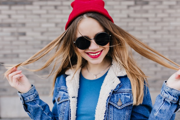 Encantadora mujer joven con maquillaje brillante pasando el día de primavera al aire libre. Foto de hermosa niña blanca en chaqueta de mezclilla riendo delante de la pared de ladrillo.