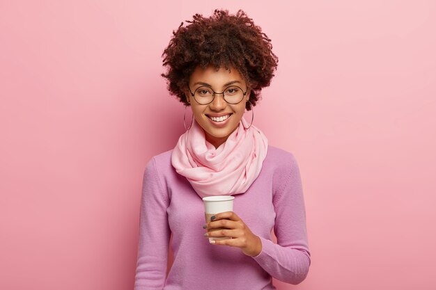 Encantadora mujer joven con peinado afro, bebe café para llevar de vaso desechable