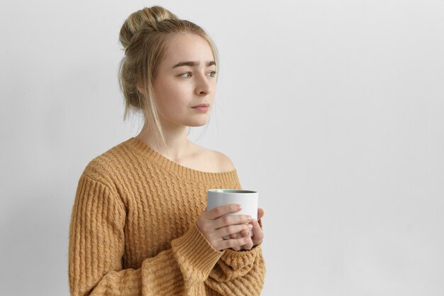 Encantadora mujer joven con peinado desordenado y jersey de punto de gran tamaño posando en una pared gris en blanco, sosteniendo una taza grande, bebiendo té, café, cacao o chocolate caliente por la mañana