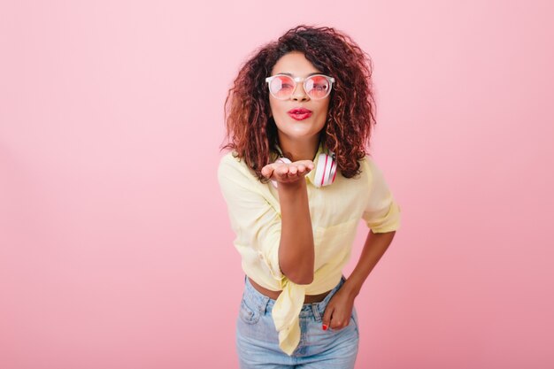 Encantadora mujer joven de pelo oscuro en ropa casual envía beso al aire. Captura de dama africana en jeans y camisa amarilla disfrutando del tiempo libre