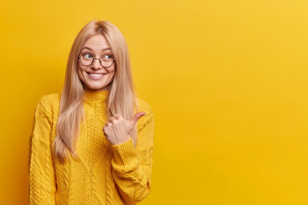 Encantadora mujer joven de pelo rubio tiene una sonrisa amistosa, apuntando al espacio de la copia en la pared amarilla