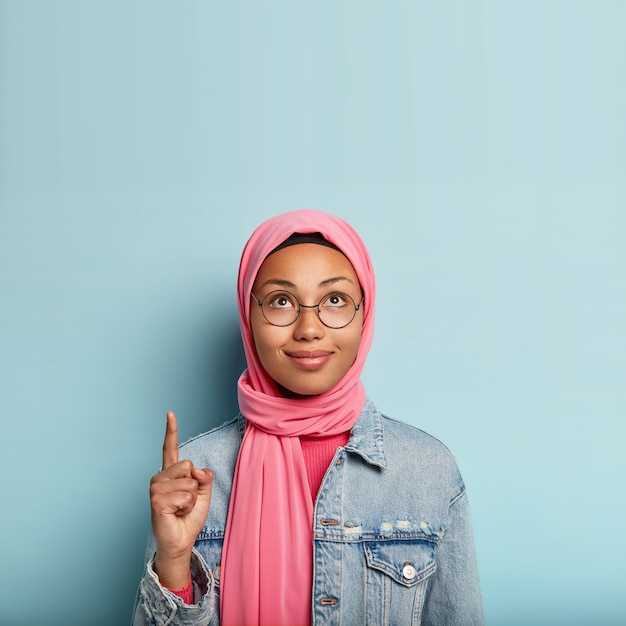 Encantadora mujer joven con piel oscura y saludable, mira y señala hacia arriba, indica con el dedo índice, usa hijab rosa, chaqueta de moda, se para sobre una pared azul, anuncia espacio de copia, muestra la manera correcta