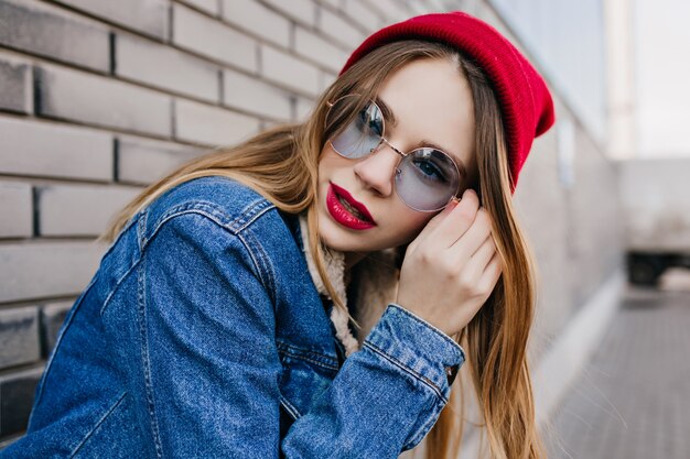 Encantadora mujer joven con piel pálida posando junto a la pared de ladrillo con expresión seria. Foto al aire libre de la chica de moda en el sombrero rojo de moda escalofriante en la calle.