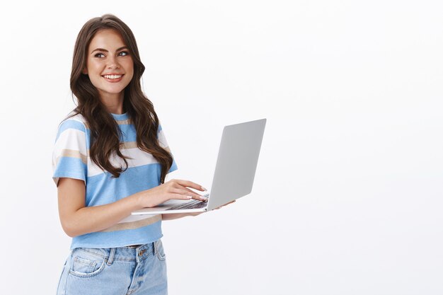 Encantadora mujer joven que trabaja por cuenta propia, alejada de la oficina, sostenga la computadora portátil, escribiendo un ensayo universitario, gire a la izquierda sonriendo alegremente, navegando por la web usando la computadora