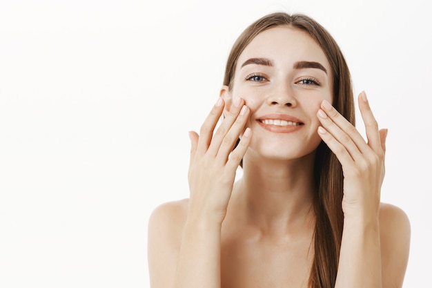 Encantadora mujer joven relajada y suave haciendo un procedimiento cosmetológico aplicando crema facial en la cara con los dedos y sonriendo ampliamente sintiéndose perfecta, cuidando la piel