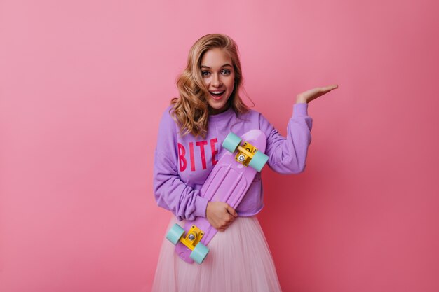 Encantadora mujer joven en ropa de moda que expresa felicidad. Chica extasiada con cabello ondulado con patineta púrpura.