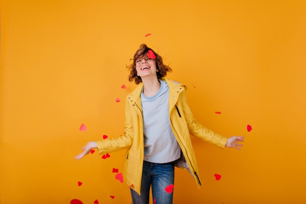 Encantadora mujer joven en ropa de otoño divirtiéndose y tirando corazones. Adorable chica en jeans preparándose para el día de san valentín.