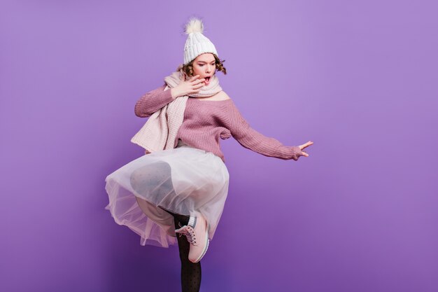 Encantadora mujer joven con sombrero gracioso posando emocionalmente en la pared púrpura. Foto de interior de una encantadora modelo femenina con una exuberante falda blanca y accesorios de invierno.