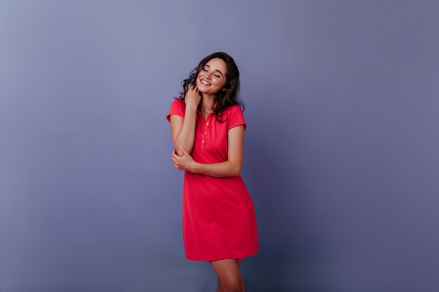 Encantadora mujer joven sonriendo con los ojos cerrados en la pared púrpura. Foto interior de alegre chica rizada en vestido rojo de moda.