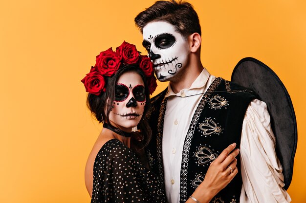 Encantadora mujer joven en traje de mascarada posando en la pared naranja. Hombre alegre en sombrero y maquillaje de halloween abrazando a su novia.