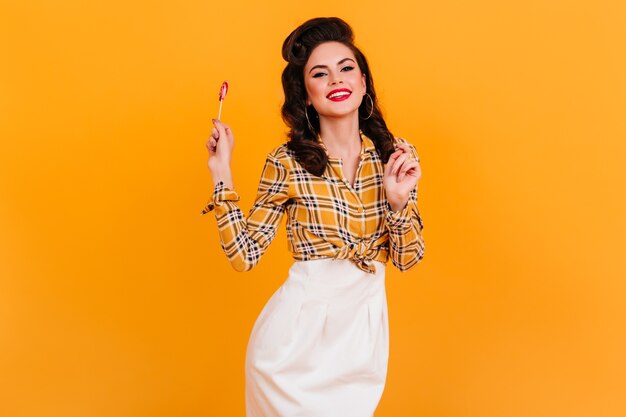 Encantadora mujer joven en traje vintage con dulces. Chica pinup sonriente con piruleta de pie sobre fondo amarillo.