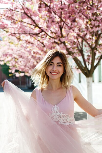 Encantadora mujer joven en vestido rosa posa ante un árbol de sakura lleno de flores rosadas