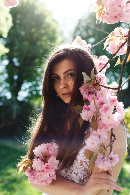 Encantadora mujer joven en vestido rosa posa ante un árbol de sakura lleno de flores rosadas